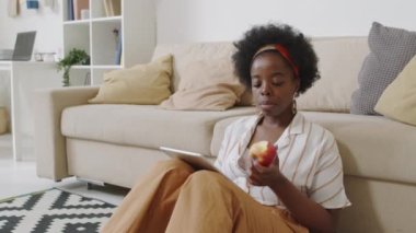 Orta yavaş çekimde, rahat bir dairedeki kanepenin yanında, dijital tablette kayarken, gündelik giysiler içinde, elma yiyen genç Afro-Amerikalı bir kadın görülüyor.