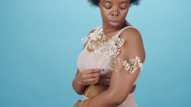 Yumuşak, genç, güzel, iri yarı, Afro-Amerikan kökenli bir kadının eğik portresi. Mavi arka planda bebek nefesli bir çiçekle iç çamaşırlarıyla poz veriyor.
