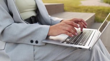 Orta bölüm, iş sırasında laptopta klavyede yazan gri pantolonlu, tanınmayan iş kadınının yavaş elleriyle yakın çekim.