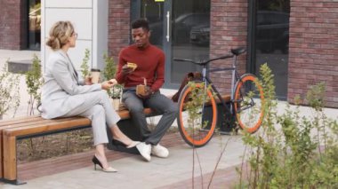 Güneşli bir günde modern ofis binasının yanındaki bankta birlikte öğle yemeği yerken çok ırklı bir çiftin sohbet edişinin yavaş çekimi.