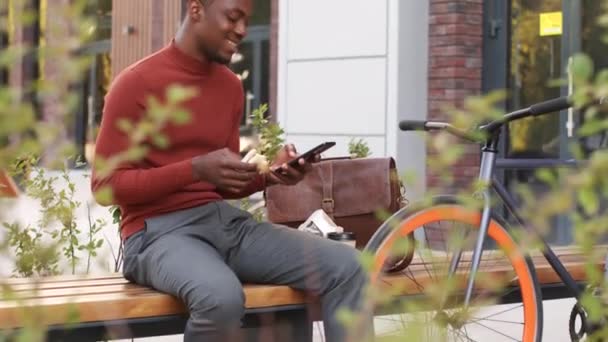 スマートなカジュアルウェアで陽気な若いアフリカ系アメリカ人の男のティルトアップの遅いショットは スマートフォンで屋外スクロールし 現代的なビジネス地区のベンチに座ってサンドイッチを食べます — ストック動画