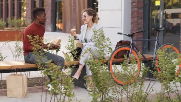 中午时分 一对年轻的多族裔同事或商业伙伴坐在市中心的长椅上聊天和吃三明治的慢镜头 — 图库视频影像