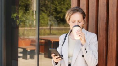Şehir merkezinde şık giyimli beyaz bir iş kadınının orta yavaş portresi. Yemek fincanından kahve içiyor, akıllı telefondan kayıyor ve kameraya gülümsüyor.