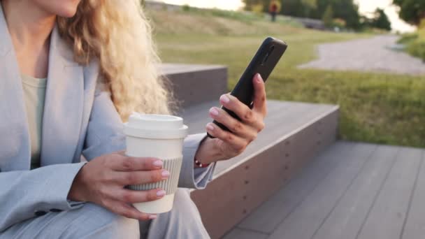 身着灰色裤子的女商人一边拿着可重复使用的咖啡杯一边在户外停车场翻看智能手机 慢吞吞地坐在中间 — 图库视频影像