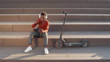 Zeki günlük giyimli ve kulaklıklı genç siyahi adamın yavaş çekimleri açık havada, e-scooter 'ın yanında oturmuş akıllı telefondan enerjik bir müzik videosu izliyor.