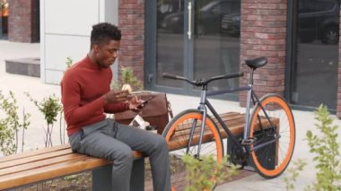 Akıllı giyimli genç Afro-Amerikan bir adamın akıllı telefondan sarkan ve şehir merkezindeki bisikletinin yanında bankta oturmuş sandviç yerken yavaş çekimleri.