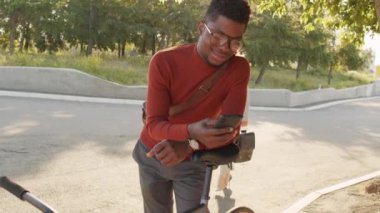 Yakışıklı Afro-Amerikan iş adamının orta yavaşlığı şık günlük kıyafetleri ve gözlükleriyle akıllı telefona sarkıyor güneşli bir günde bisiklet koltuğuna yaslanıyor.