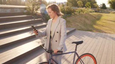 Güneşli bir günde şehir parkında bisiklet sürerken akıllı günlük giysiler içinde akıllı telefon kullanan gülümseyen genç iş kadınının orta yavaş portresi.