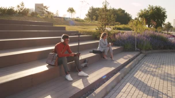在阳光灿烂的日子里 年轻的非洲裔美国男人拿着电动车在耳机里欣赏音乐 而白种人的女商人则在智能手机上打滚 坐在公园的楼梯上 — 图库视频影像