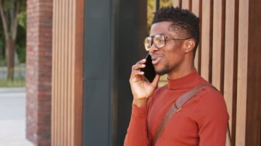 Güneşli bir günde şehrin göbeğinde akıllı telefondan sohbet ederken gülümseyen Afro-Amerikan bir adamın yavaş çekimleri.