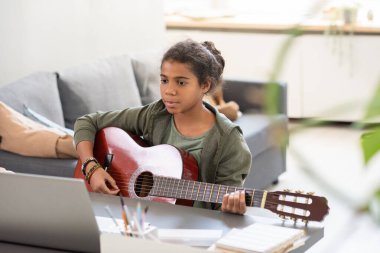 Dizüstü bilgisayarının önünde gitar çalan ciddi bir kız öğrenci.