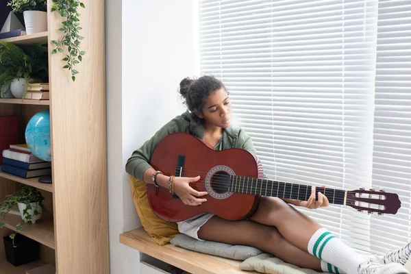 Rilassato studentessa interculturale suonare la chitarra da finestra — Foto Stock