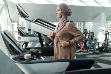 Genç bayan tesettürlü ve aktif giyimli spor makinelerinden birinde egzersiz yapıyor.