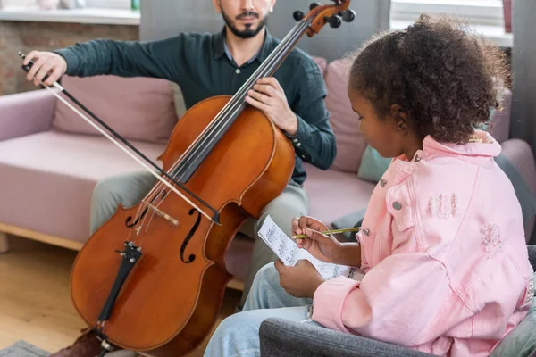 Menina intercultural escrevendo notas musicais em papel durante a aula de violoncelo — Fotografia de Stock