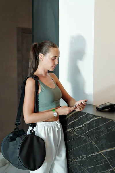 Brunetka dziewczyna w ubraniu aktywnym patrząc na ekran smartfona stojąc przy recepcji — Zdjęcie stockowe