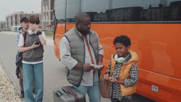 アフリカ系アメリカ人の父親と息子の中程度の手持ち撮影で オレンジバスの隣に立つ乗客が運転手を待っているチケットを手に持っている — ストック動画