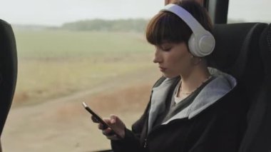 Gündelik kıyafet ve kablosuz kulaklık takan kısa saçlı kız öğrencilerin orta boy yakın çekimi otobüs yolculuğu sırasında pencerenin kenarında oturmuş akıllı telefondan kayıyor.