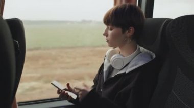 Kısa saçlı, günlük kıyafetli, kablosuz kulaklık takan ve otobüs yolculuğu sırasında manzaraya bakan orta sınıf bir kız çocuğu.