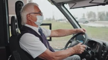 Şık üniformalı ve yüz maskeli yaşlı otobüs şoförünün orta boy portresi sürücü koltuğunda direksiyon tutarken kameraya bakıyor.