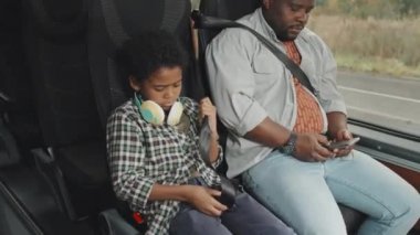 Otobüsten önce babasıyla birlikte otobüste otururken, günlük giyimli, sevimli, Afro-Amerikan bir çocuğun orta boy fotoğrafı. Akıllı telefondan mesaj atan adam