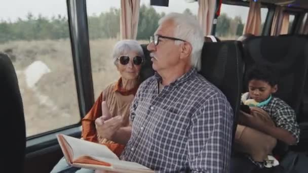 会話をしながらバスの中で一緒に座って幸せな高齢者のカップルの中ショット 男は紙の本を持ちながら窓のそばに座って妻と話していた — ストック動画