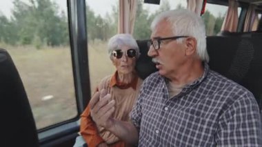 Orta boy beyaz giysili ve gözlüklü bir adamın otobüste birlikte otururken karısıyla konuştuğu fotoğraf.