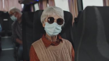 Otobüste oturan, tıbbi maske takan, koyu renk güneş gözlüklü, ciddi bir son sınıf öğrencisinin orta boy portresi.