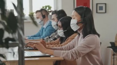 Yüz maskesi ve kulaklık takmış Asyalı bir kadının portresi müşteriyle konuşuyor ve çağrı merkezinde bilgisayarla çalışıyor, sonra da kameraya bakıyor.