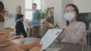 Yüz maskeli ve gözlüklü Asyalı bir kadının pandemik görüşme sırasında anti-damlacık plastik ekranın arkasında oturduğu ve İK yöneticileriyle konuştuğu orta boy bir fotoğraf.