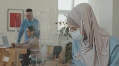 Müslüman kadının tesettür ve yüz maskesi takıp koruyucu ekranın arkasında oturduğu ve salgın sırasında başarılı bir toplantı veya iş görüşmesi yaptıktan sonra şarkı söylediği sahneler.