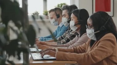 Çağrı merkezi operatörlerinin yüz maskeli görüntülerinin izlenmesi kulaklık ve dizüstü bilgisayar kullanarak ve ofisteki müşterilere yardım ederek.