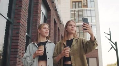 Küçük ikiz kız kardeşlerin, kahve bardaklarına kahve doldurup sokakta yürürken cep telefonlarında gülüşlerini ve videolarını çektikleri düşük açılı bir görüntü.