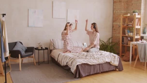 Pan拍摄到一对快乐的孪生姐妹坐在床上 用耳机听着音乐跳舞 — 图库视频影像
