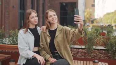 Dışarıda bankta oturan ve cep telefonuyla selfie çeken güzel ikiz kız kardeşlerin fotoğrafını çekiyorum.