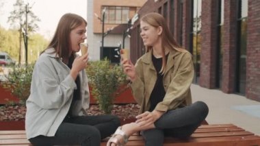 Dışarıda bankta oturmuş dondurma yerken sohbet eden mutlu ikiz kız kardeşlerin orta boy fotoğrafı.