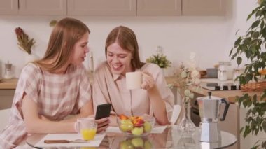 Genç ve mutlu ikiz kız kardeşlerin rahat bir mutfakta oturmuş kahve içerken sohbet ederken ve cep telefonuyla sosyal medyada gezinirken PAN fotoğrafı.