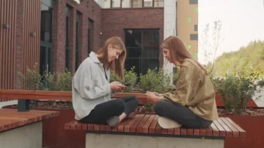 Dışarıda bankta bacakları bağlı oturan ve sosyal medyada cep telefonuyla sohbet eden genç ikiz kız kardeşlerin görüntülerini izliyoruz.