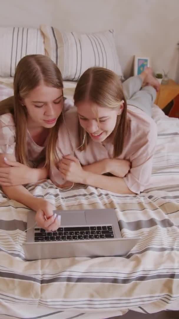 ラップトップを使用している間にベッドの上に横たわって笑っている同じ双子の姉妹の垂直ショットを開催 — ストック動画