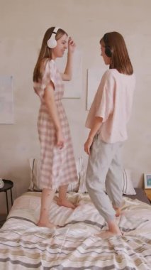 Evde yatakta kulaklık takıp dans eden mutlu ikiz kız kardeşlerin el kamerasıyla çekilmiş resmi.