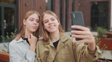Dışarıda bankta oturup cep telefonuyla selfie çeken mutlu ikiz kız kardeşlerin el kamerasıyla çekilmiş görüntüleri.