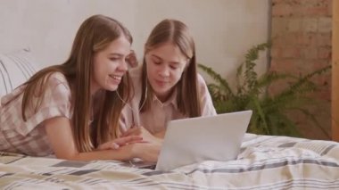 Yatakta uzanmış kulaklıkla müzik dinlerken ve dizüstü bilgisayar kullanarak sohbet eden mutlu ikiz kız kardeşlerin PAN çekimi.