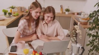 Mutlu ikiz kız kardeşlerin pastel renkli giysiler giyip rahat bir mutfakta oturmuş sohbet ederken ve dizüstü bilgisayar kullanırken gülüşlerinin el kamerası görüntüsü.