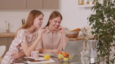 Genç ve mutlu ikiz kız kardeşlerin rahat bir mutfakta oturmuş bilgisayardan biriyle sohbet ederken PAN fotoğrafı.