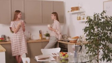 Pastel renkli elbiseli genç ikiz kız kardeşlerin sabah kahvenin tadını çıkarırken rahat bir mutfakta durup sohbet etmelerinin PAN fotoğrafı.