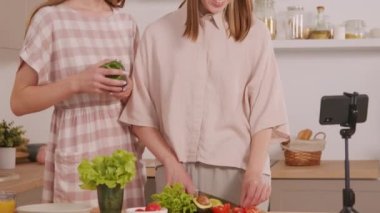 Neşeli, genç ikiz kız kardeşlerin sebze keserken ve rahat mutfakta salata hazırlarken cep telefonundan yemek videosu çekimleri.