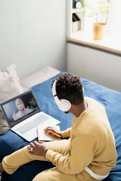 在网上上课的时候，戴着耳机、身穿家居服的年轻学生坐在床上 — 图库照片
