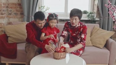 Üç kişilik Çinli bir ailenin geleneksel kırmızı kıyafetler giyip evdeki kanepede otururken ve Ay Yeni Yılı için hediye sepeti hazırlarken orta boy bir fotoğrafı.