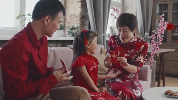幸せなアジアの両親がソファに座って 伝統的な衣装でかわいい5歳の娘に中国の旧正月のポストカードを与えることのスローモーションで撮影 — ストック動画