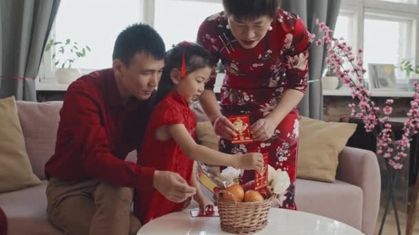 中国の旧正月のお祝いのためのギフトバスケットを準備するかわいい5歳の女の子を助ける赤いシャツの伝統的なドレスと父でアジアの母の中のショット — ストック動画