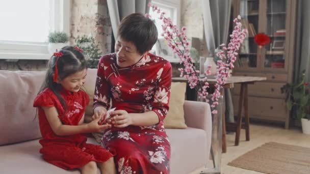 中国の旧正月のために飾られたリビングルームでソファに座ってかわいい5歳の女の子に赤いポストカードを与える伝統的なドレスで幸せなアジアの母親のスローモーションで撮影 — ストック動画